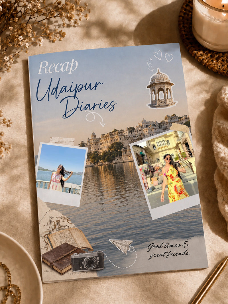 A custom photo collage journal titled 'Recap Udaipur Diaries' displayed on a beige fabric surface with styled props like dried flowers, a candle, and a book. The cover features a scenic lake view of Udaipur, two polaroid-style photos of a woman, a camera, and handwritten text.