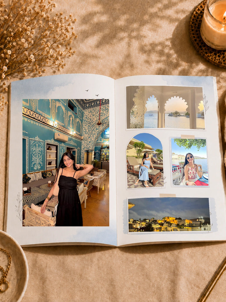 An open custom photo album displayed on a beige fabric surface, featuring printed travel photographs of a woman in a black dress in an ornate blue interior, white archways by water, and a nighttime city view. A lit candle, dried flowers, and a bowl are styled around the album.