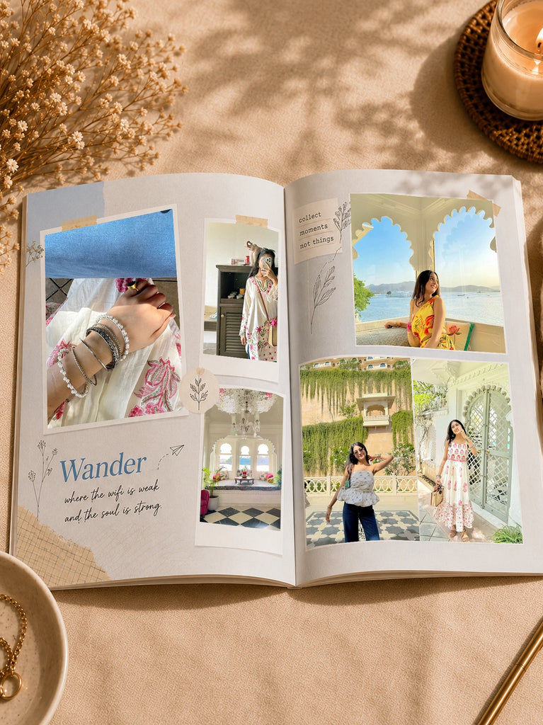 An open custom photo album spread on a beige fabric surface, surrounded by dried flowers, a lit candle, and a bowl of jewelry. The pages feature printed layouts with embedded photos of women in traditional and casual outfits, scenic backgrounds, and decorative text including 'Wander'.